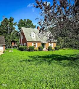 A red brick house with a green roof sits on a large, grassy lawn. The house has two dormer windows, a chimney, and shrubs lining the front. Trees are visible in the background under a clear blue sky.