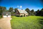 A two-story brick house with white trim and a steep gray roof sits beside a detached two-car garage at 8683 US Highway 15 in Bullock. The property features a long driveway, green lawn, and trees in the background under a blue sky.