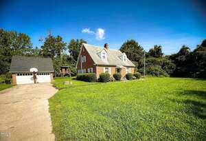 A two-story brick house with white trim and a steep gray roof sits beside a detached two-car garage at 8683 US Highway 15 in Bullock. The property features a long driveway, green lawn, and trees in the background under a blue sky.