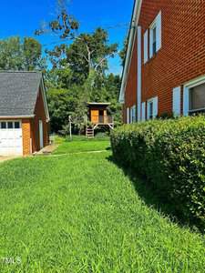 A green lawn runs between a brick house and a garage. In the background, a small wooden structure on stilts with stairs is visible, surrounded by trees and shrubs under a clear blue sky.