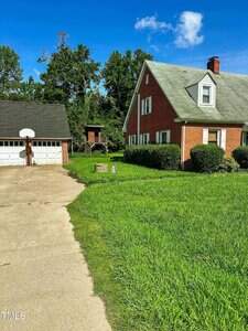 A red brick house with a gray roof sits beside a two-car garage with a basketball hoop. The driveway curves past a grassy yard, with trees and a wooden playset in the background under a blue sky.