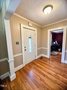 A hallway with light tan walls, white trim, and hardwood floors features a white front door with a decorative glass panel. An adjacent room with dark red walls and a piano is visible through a doorway. Ceiling light fixture is on.