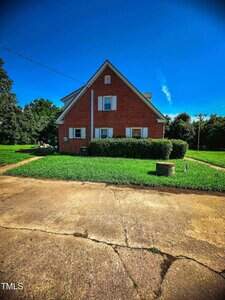A red brick house with a steep gable roof and white trim sits on a grassy lawn. A concrete driveway with visible cracks is in the foreground, and the sky is clear and blue.