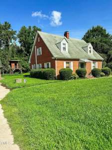 A two-story brick house with a green roof, white trim, and dormer windows sits behind a small porch and neatly trimmed bushes, surrounded by a large lawn and trees under a bright blue sky.