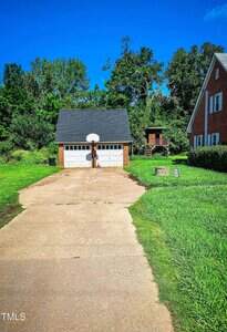 A large concrete driveway leads to a detached two-car garage with a basketball hoop above the doors. There is a grassy yard, trees in the background, and part of a brick house visible on the right.