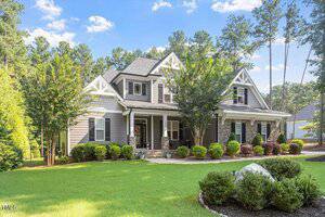 A two-story gray house with white trim, a covered front porch supported by stone columns, surrounded by neatly trimmed bushes, trees, and a well-maintained lawn under a blue sky.