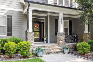 A covered front porch with stone pillars, black rocking chairs, potted plants, and a black front door with a wreath. The house exterior is gray with white trim and black shutters. Shrubs and mulch border the walkway.