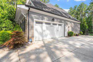 A modern suburban house garage with gray siding, a large white double garage door, small windows above the door, stone detailing on one corner, and landscaped shrubs along the side. The driveway is concrete.