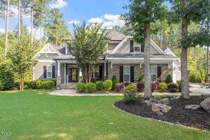 Nestled in Wake Forest at 1203 Smith Creek Way, this two-story suburban house features gray siding, stone accents, and white trim amid green grass, trees, and landscaped bushes, set against a backdrop of tall pines under a blue sky.