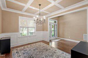 A bright, empty dining room with tan walls, white wainscoting, a coffered ceiling, a chandelier, hardwood floors, a patterned area rug, a window, and glass double doors leading outside.