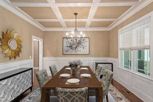 A dining room with a dark wood table set for six, upholstered chairs, a modern chandelier, wainscoted walls, a large window with blinds, and contemporary artwork and mirror on the walls.
