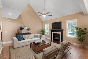 A spacious living room with vaulted ceiling, tan walls, hardwood floors, a white sofa, two accent chairs, a black fireplace with a mounted TV, a sideboard, houseplants, and large windows letting in natural light.