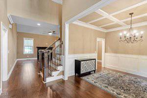 A spacious entryway with hardwood floors, beige walls, white wainscoting, a staircase with dark railings, a modern sideboard, a chandelier, and a patterned rug under a coffered ceiling.