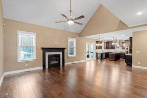 Spacious open-concept living room with hardwood floors, a fireplace, vaulted ceiling with a ceiling fan, two windows, and a view into a modern kitchen with dark cabinets and an island.