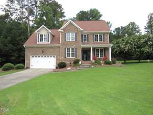 This charming two-story brick house at 1601 Carriage Drive, Franklinton, features a red shingle roof, white window frames, and a two-car garage on a large, neatly manicured lawn with shrubs and trees in the background.