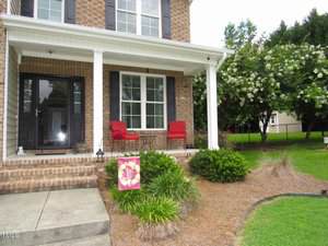 A brick house with a covered front porch featuring two red chairs and a small table. A flowering tree and well-maintained shrubs are in the yard, and a colorful flag hangs by the porch steps.