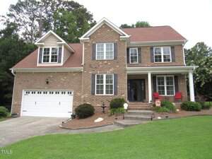 Charming two-story brick house in Franklinton with white-framed windows, a red shingled roof, attached two-car garage, front porch with red chairs, and a well-maintained lawn at 1601 Carriage Drive bordered by trees.
