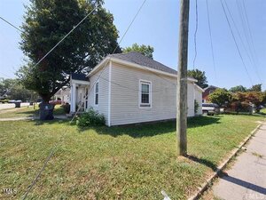 A small, single-story beige house with white trim sits on a grassy corner lot. Power lines and a utility pole are visible in the foreground, with a large tree and neighboring homes in the background on a sunny day.