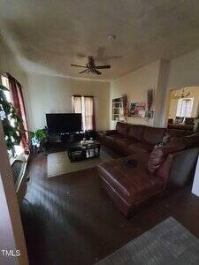 A living room with a brown sectional sofa, a coffee table, a large TV on a stand, a ceiling fan, and several windows with curtains. There are plants and shelves with books and decorative items.