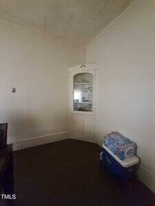 A corner of a sparsely furnished room with white walls, a built-in white corner cabinet containing a few items, and a blue cooler topped with a pack of bottled water on the dark floor.