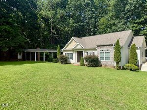 Single-story house with beige siding, stone accents, and multiple front gables. The home has a large grassy lawn, manicured bushes, a covered carport on the left, and is surrounded by tall trees in the background.