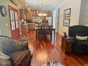 A cozy living and dining area with wood flooring, brown furniture, a dining table with four chairs, an armchair, a patterned chair, and a view into a kitchen with wooden cabinets and white appliances.