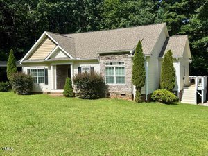 Charming single-story house at 81 White Deer Lane, Henderson, featuring a beige and stone exterior, gable roof, and multiple windows. Enjoy the lush front yard with shrubs and trees, a grassy lawn, wooded backdrop, and steps to a side entrance.