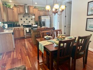 A kitchen and dining area with wooden floors, light wood cabinets, stainless steel appliances, a rectangular table set for four, and a chandelier overhead. The kitchen features a range hood and tile backsplash.
