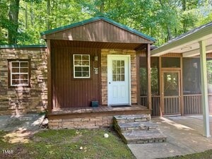 Small wooden house with a covered front porch, stone accents on the exterior, a white door, two windows, and an attached screened-in porch, surrounded by trees and greenery.