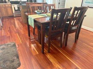 A wooden dining table with four chairs is set with dishes and placemats on a polished hardwood floor in a kitchen and dining area. A green table runner and area rug are also visible.