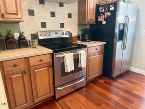 A kitchen with wooden cabinets, stainless steel oven, two white towels on the oven handle, a black refrigerator, coffee maker, and decorative jars on the countertop. The backsplash features a checkerboard tile pattern.