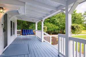 A covered porch with light blue wooden flooring, white railings, and a blue cushioned porch swing. Trees and greenery are visible in the background.