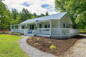 A white single-story house with a blue metal roof and wraparound porch sits amid green grass, landscaped mulch beds, and tall trees under a partly cloudy sky. A gravel path leads to the front steps.