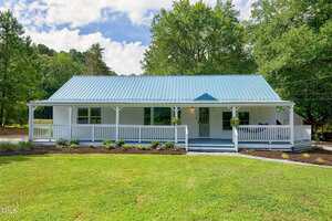 A single-story house with a blue metal roof, white siding, and a wide covered front porch. The porch is bordered by white railings, and the house is surrounded by green grass and trees.