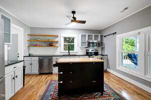 Modern kitchen with gray cabinets, stainless steel appliances, open wooden shelves, a central black island with drawers, hardwood floors, a colorful rug, and large windows letting in natural light.