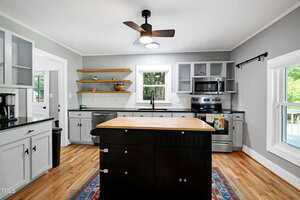 Modern kitchen with gray cabinets, black island, stainless steel appliances, wooden shelves, and light wood flooring. A window above the sink and another on the right bring in natural light. Ceiling fan and recessed lights are visible.