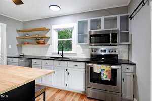 Modern kitchen with gray cabinets, stainless steel appliances, open wood shelves, a black faucet, white subway tile backsplash, and a window above the sink. A towel with an abstract design hangs on the oven handle.