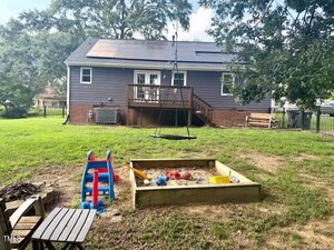 A backyard with a sandbox filled with toys, a small red and blue slide, a swing hanging from a tree, a wooden deck with stairs, and a gray house with solar panels on the roof in the background.