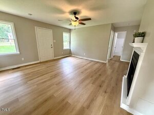 Unfurnished living room with light wood flooring, beige walls, a ceiling fan with light, two windows, a white front door, a white fireplace, and a hallway leading to other rooms. Natural light enters from the windows.