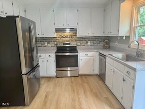 A kitchen with white cabinets, stainless steel appliances including a refrigerator, oven, and dishwasher, light wood flooring, a gray brick backsplash, and a "Good Eats" sign above the stove.