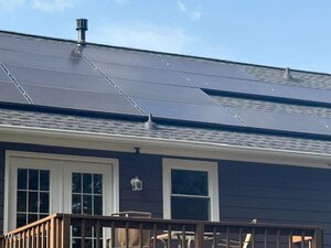 A close-up view of a house roof with multiple solar panels installed, above a deck with chairs and a table; the exterior wall is brown with white-trimmed windows and doors.