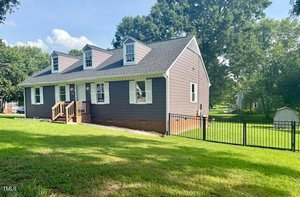 A gray house with white trim, three dormer windows, and a small front porch sits on a grassy lawn. A black metal fence encloses part of the yard, and there is a shed in the background. Trees and blue sky are visible.