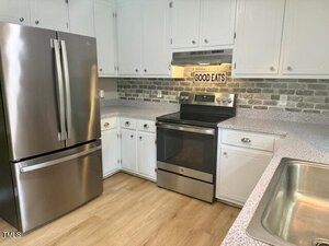 A modern kitchen with white cabinets, stainless steel refrigerator and oven, a brick-patterned backsplash, light-colored countertops, a double sink, and a sign above the stove that reads "GOOD EATS.