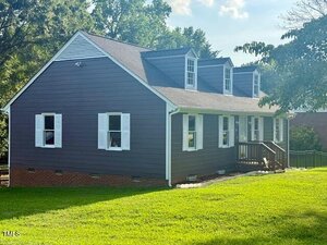 A dark gray, single-story house with white trim and dormer windows sits on a brick foundation. A small wooden porch leads to the front door, and the house is surrounded by green grass and trees under a sunny sky.
