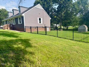 A gray two-story house with white trim sits on a grassy lawn. There is a black metal fence enclosing part of the yard and a small storage shed in the background surrounded by trees.