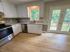 A kitchen with white cabinets, stainless steel appliances, light wood flooring, a window above the sink, and glass double doors opening to a deck with a view of green trees.