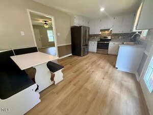 A kitchen with light wood flooring, white cabinets, stainless steel appliances, and a black refrigerator. A white breakfast nook with black cushions sits in the left corner. A doorway leads to another room.