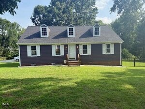 A dark gray, one-story house with three dormer windows on the roof, white trim, and a small front porch with steps, set on a grassy lawn with trees in the background.