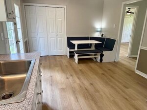 A kitchen with a stainless steel sink in the foreground, light wood flooring, white cabinets, a built-in corner bench with a table, and a small lamp on the table. White double doors are on the back wall.