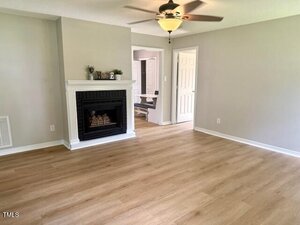 A living room with light wood flooring, a white fireplace with a black insert, beige walls, a ceiling fan with lights, and an open doorway leading to another room.